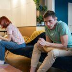 Jealous man husband sitting on sofa with wife typing message on smartphone