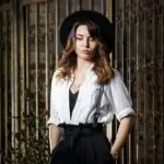 Young woman in white shirt and black hat standing near the gate.