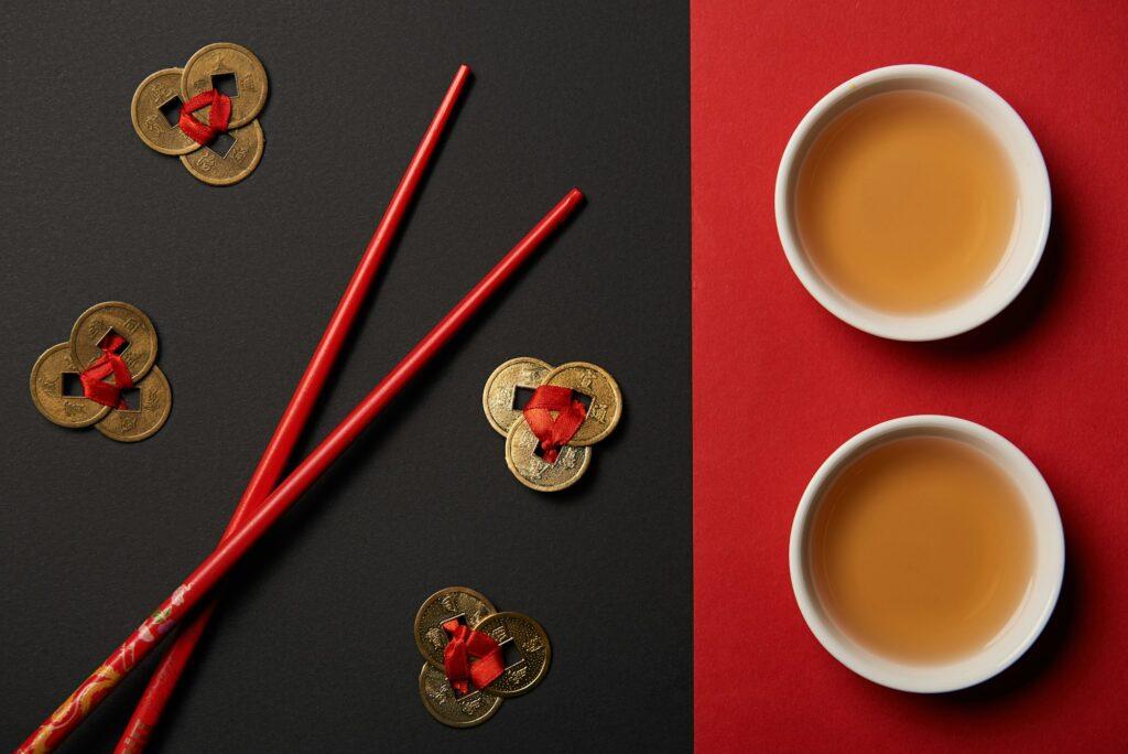 top view of chopsticks with traditional chinese tea and feng shui coins on red and black background