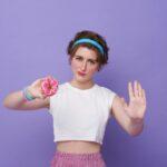 Young sad woman hold eat burger show hand stop gesture say no junk food