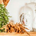 A serene still life featuring a bust with lipstick traces, boxwood, dried grass, and a glass carafe.