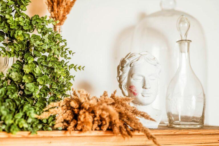 A serene still life featuring a bust with lipstick traces, boxwood, dried grass, and a glass carafe.
