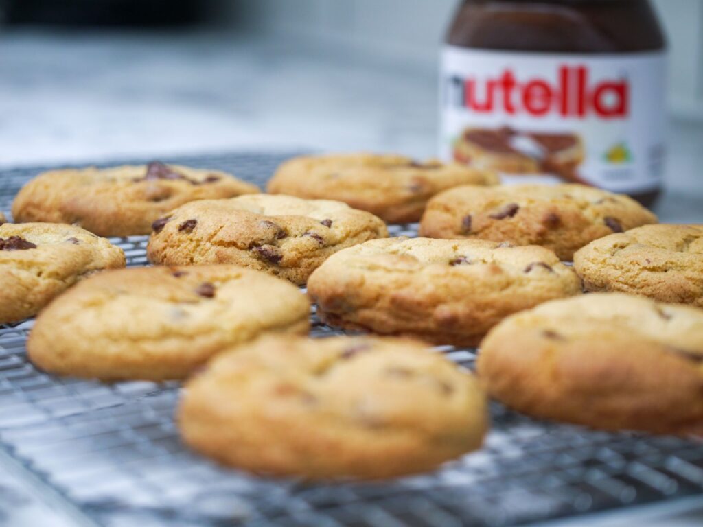 Gros plan sur des cookies aux pépites de chocolat faits maison refroidissant avec un pot de Nutella en arrière-plan.