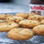 Gros plan sur des cookies aux pépites de chocolat faits maison refroidissant avec un pot de Nutella en arrière-plan.