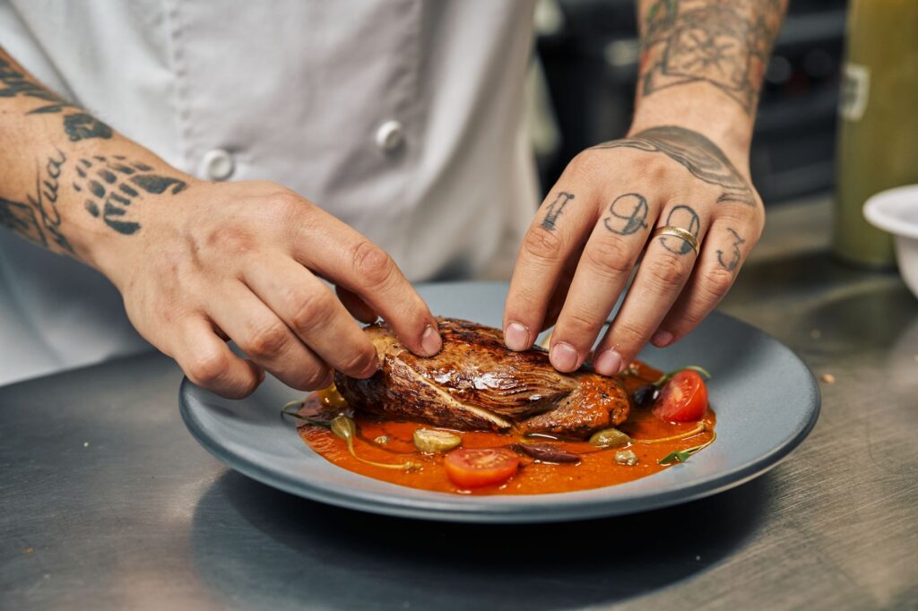 Un chef tatoué présente un steak parfaitement cuit, garni de divers accompagnements, sur une assiette colorée.