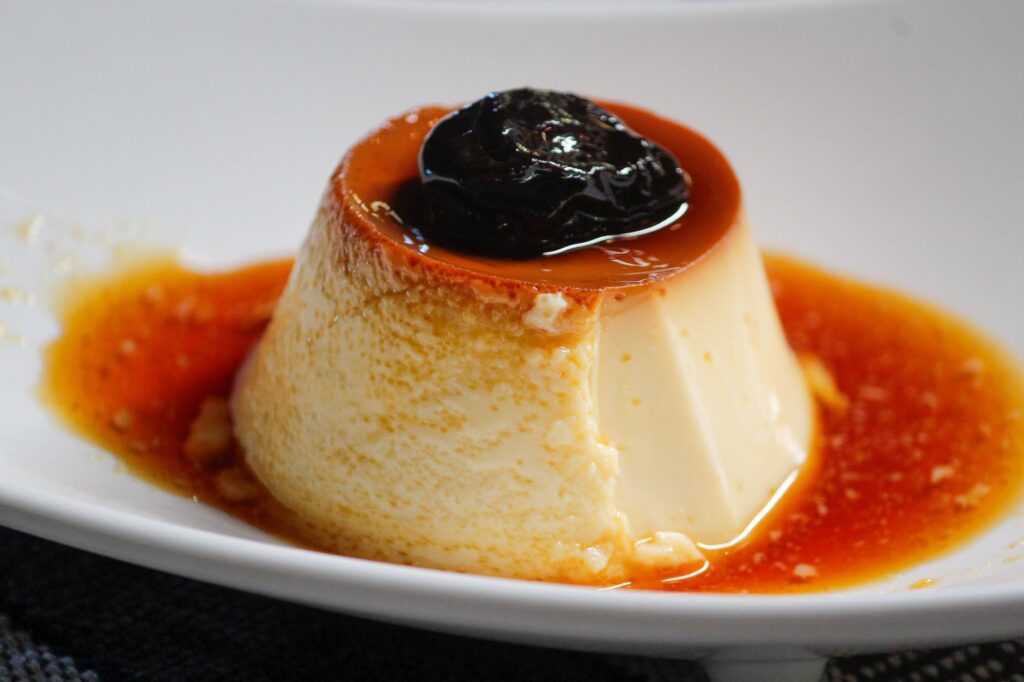 Flan crémeux nappé de sauce caramel et garni d'un pruneau, servi sur une assiette blanche.