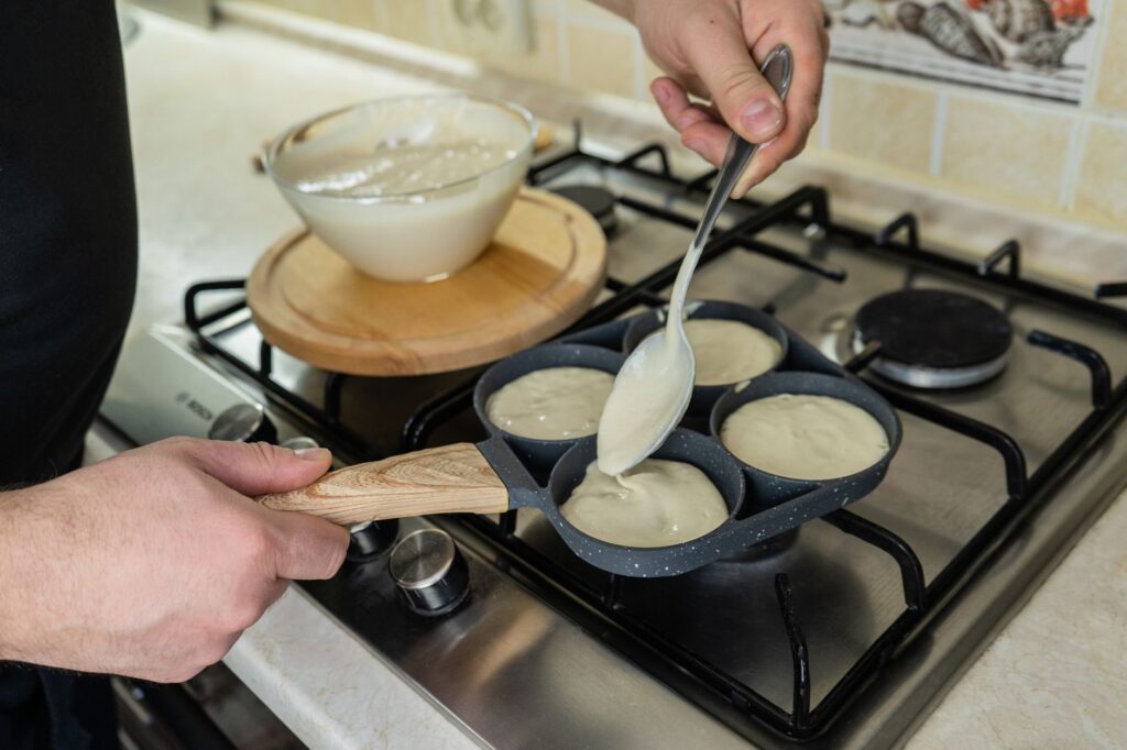 Gros plan sur une personne préparant des crêpes sur une plaque de cuisson à l'aide d'une louche et d'une poêle remplie de pâte.