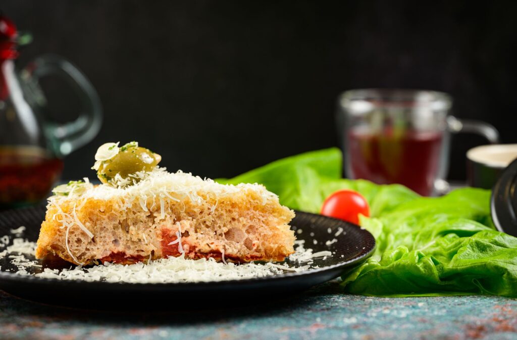 Une délicieuse tranche de focaccia garnie de fromage et d'olives sur une assiette noire, décorée de légumes frais.