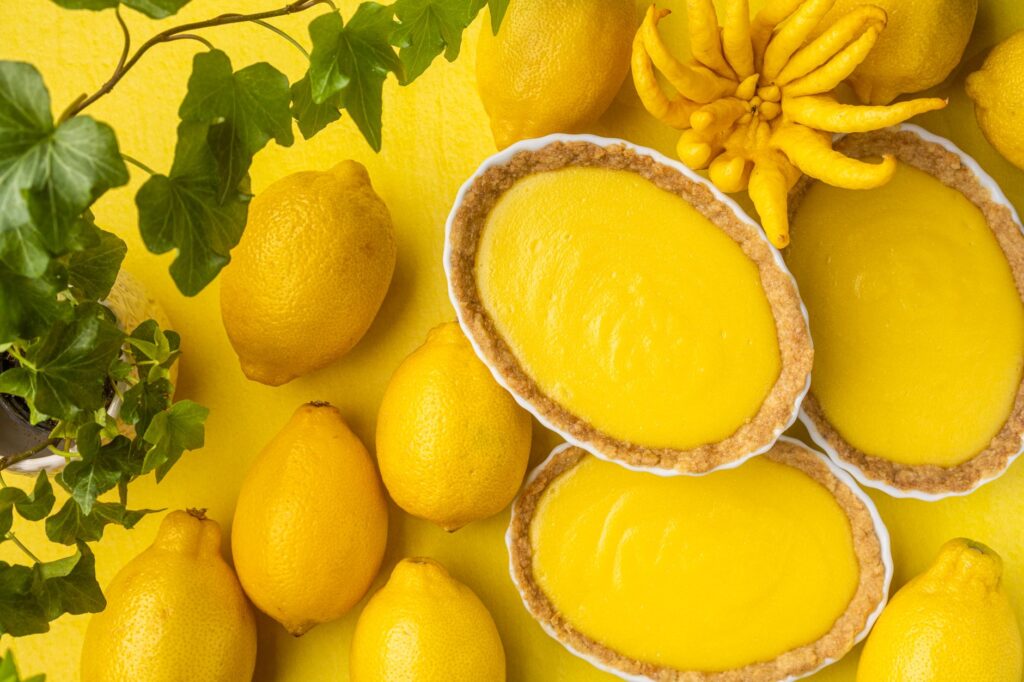 Des tartes au citron lumineuses et appétissantes entourées de citrons frais et de verdure sur un fond jaune.