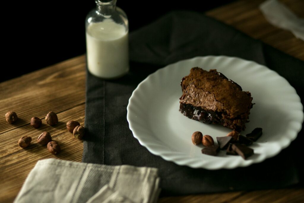 Une riche part de gâteau au chocolat sur une assiette blanche avec des noisettes et des morceaux de chocolat sur une table en bois rustique.