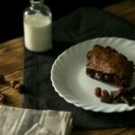 Une riche part de gâteau au chocolat sur une assiette blanche avec des noisettes et des morceaux de chocolat sur une table en bois rustique.