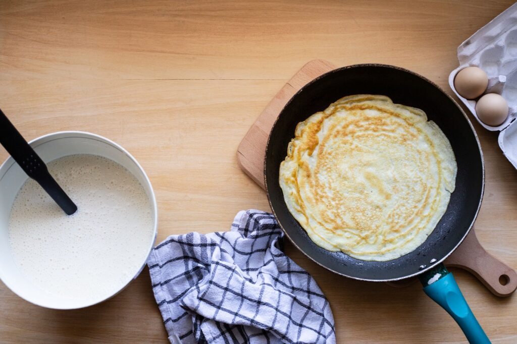 Vue de dessus d'une crêpe cuisinée dans une poêle avec de la pâte et des œufs à proximité.