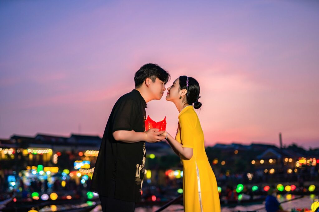 Un couple s'embrasse doucement, tenant une lanterne rouge sous un coucher de soleil coloré au bord de l'eau vibrant.