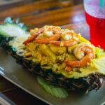 Délicieux riz frit thaïlandais à l'ananas avec des crevettes servi dans une coque d'ananas avec une boisson gazeuse rouge à côté.