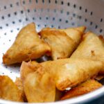 Des samosas frits croustillants et dorés placés dans une passoire en acier inoxydable, parfaits pour un en-cas salé.
