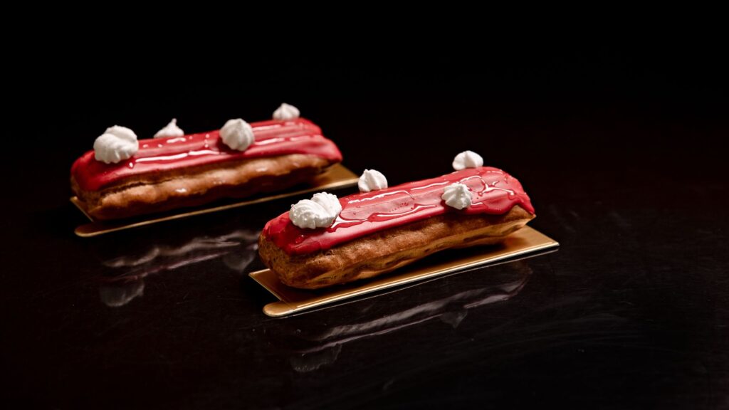 Éclairs rouges enrobés de glaçage avec décoration en crème blanche, présentés artistiquement sur une surface sombre.