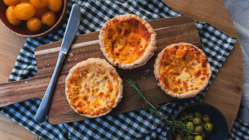Trois mini quiches fraîchement cuites sur une planche à découper avec garniture et ingrédients colorés.
