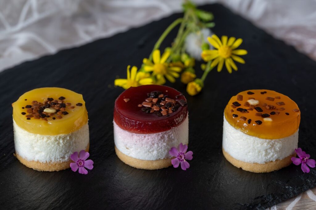 Régalez-vous de mini cheesecakes aux fruits colorés, joliment garnis de fleurs sur un plateau en ardoise foncée.