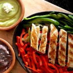 Fajitas de poulet grillé grésillant avec poivrons, guacamole et haricots frits sur une planche de bois.