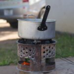 Une casserole en métal est posée sur un poêle à charbon rustique en extérieur, créant une scène de cuisine traditionnelle.