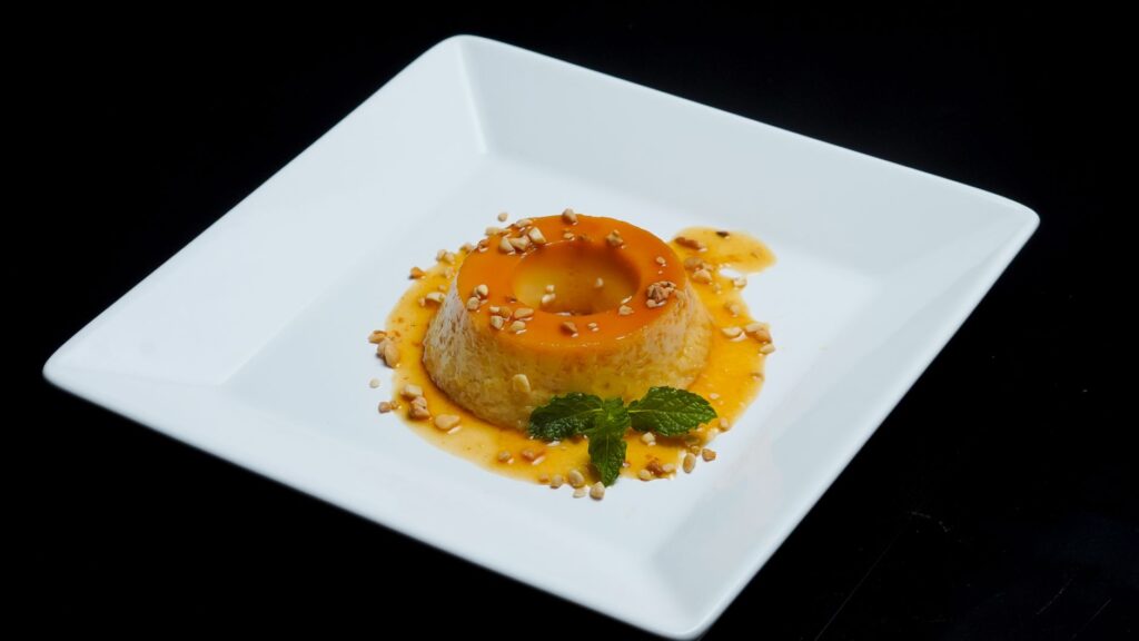 Un flan au caramel joliment présenté avec des feuilles de menthe et des noix, sur un fond noir.