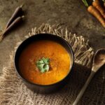 Soupe de carottes tiède dans un bol noir, garnie de persil sur un tapis texturé.