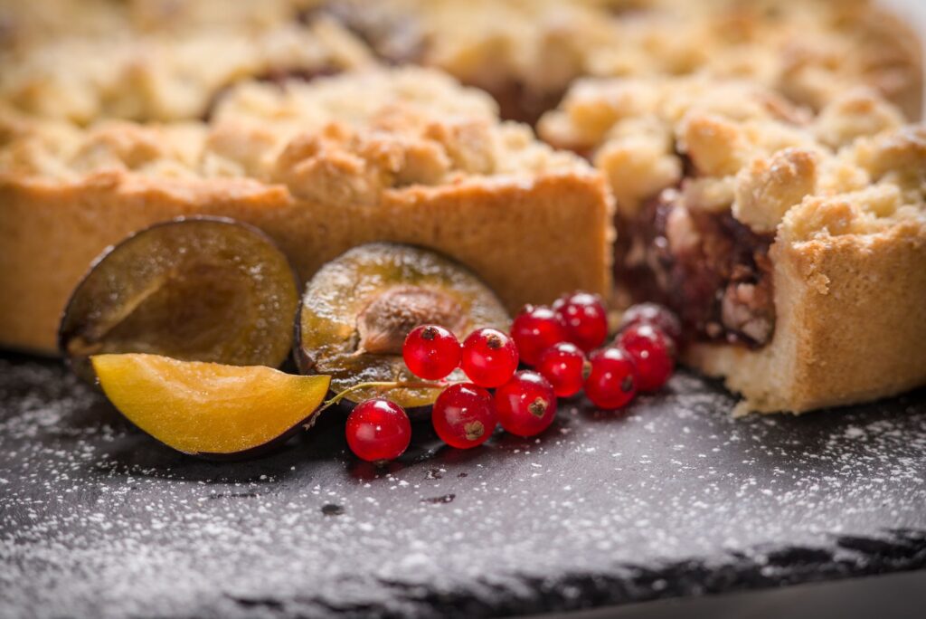 Gros plan sur une tarte crumble aux prunes avec des groseilles rouges, saupoudrée de sucre glace.