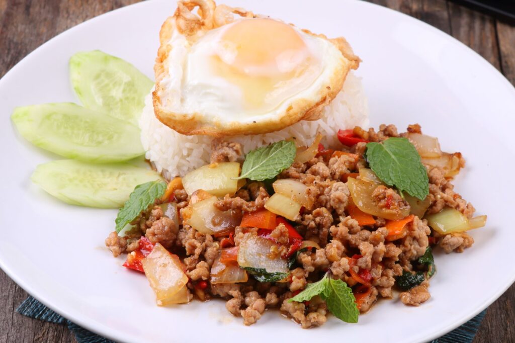 Porc au basilic thaï authentique, servi avec du riz, un œuf au plat et des concombres en garniture sur une assiette blanche.