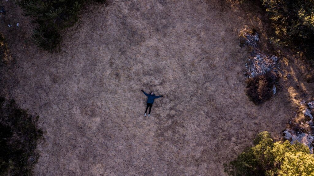 Une personne est allongée sur un champ sec entouré d'arbres, créant un sentiment de solitude vu d'en haut.
