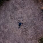 Une personne est allongée sur un champ sec entouré d'arbres, créant un sentiment de solitude vu d'en haut.