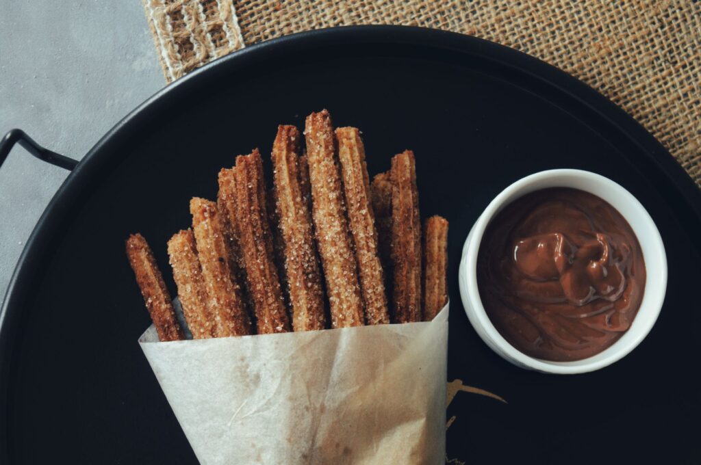 Plateau de churros emballés dans du papier, accompagné d'un bol de sauce au chocolat sur une assiette foncée.