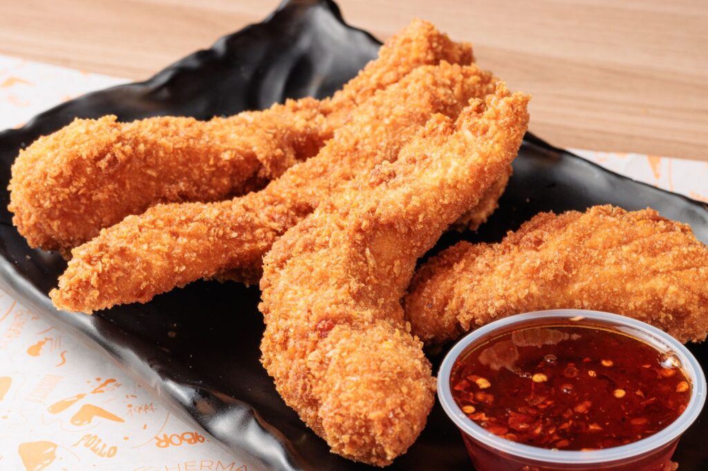 Lanières de poulet frites, dorées à souhait, servies avec une sauce chili épicée sur une assiette noire.