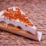 Une part alléchante de tarte banoffee garnie de crème et de caramel, posée sur un tapis en osier.