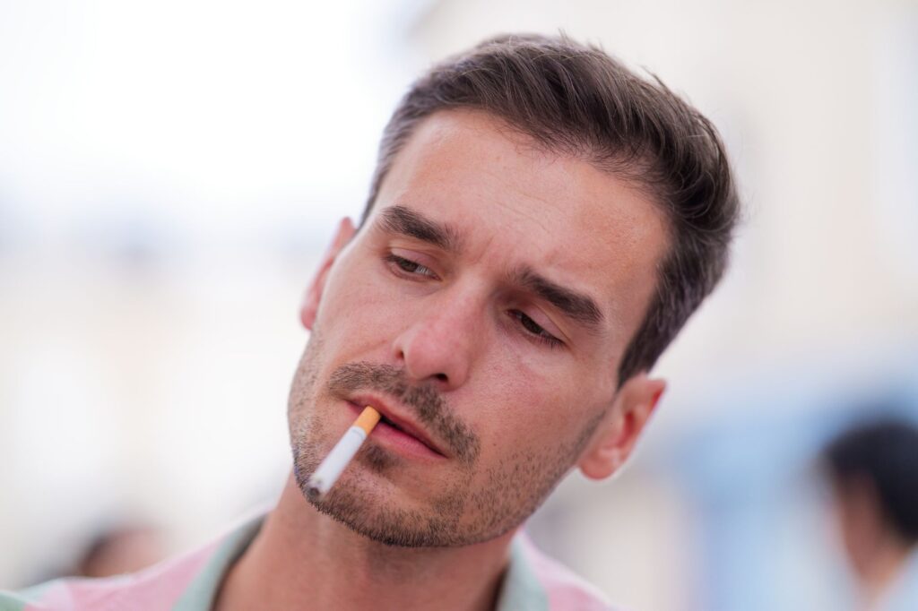 Gros plan sur un homme fumant une cigarette en extérieur, affichant une expression pensive et une ambiance cinématographique.