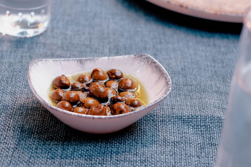 Gros plan sur des olives fraîches disposées dans un plat sur un tissu bleu texturé, évoquant la cuisine méditerranéenne.