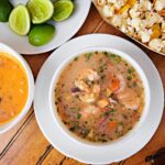 Un bol coloré de ceviche équatorien accompagné de citrons verts, de noix et de pop-corn, servi sur une table en bois.
