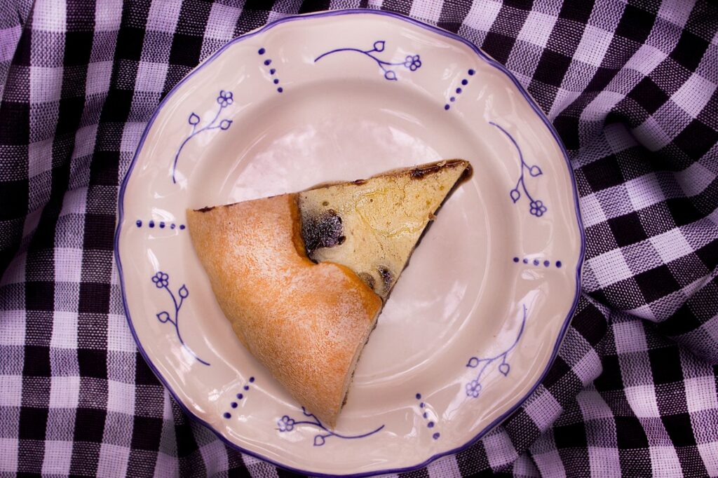 Une simple tranche de pâtisserie belge traditionnelle sur une assiette décorée de fleurs, elle-même posée sur une nappe à carreaux.