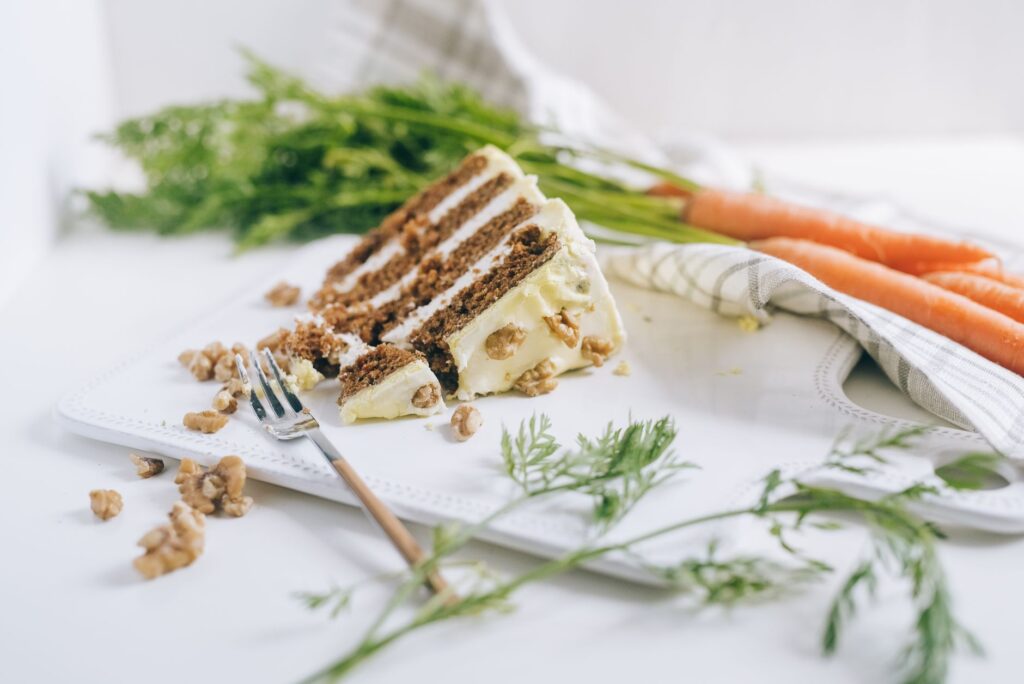 Une tranche de gâteau aux carottes avec du glaçage et des noix sur une planche à découper avec des carottes et des légumes.