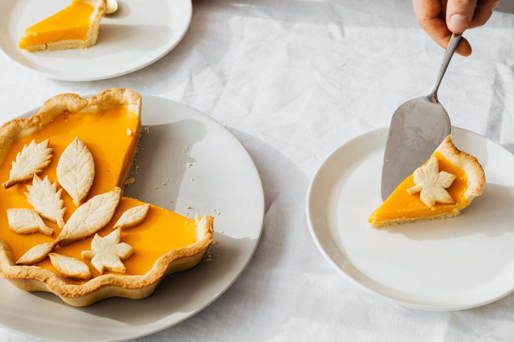 Gros plan sur une main servant une part de tarte à la citrouille avec des décorations en feuilles sur une table blanche.