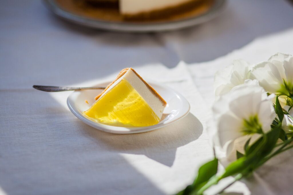 Part de cheesecake au citron sur une assiette avec des fleurs blanches sur une table ensoleillée.