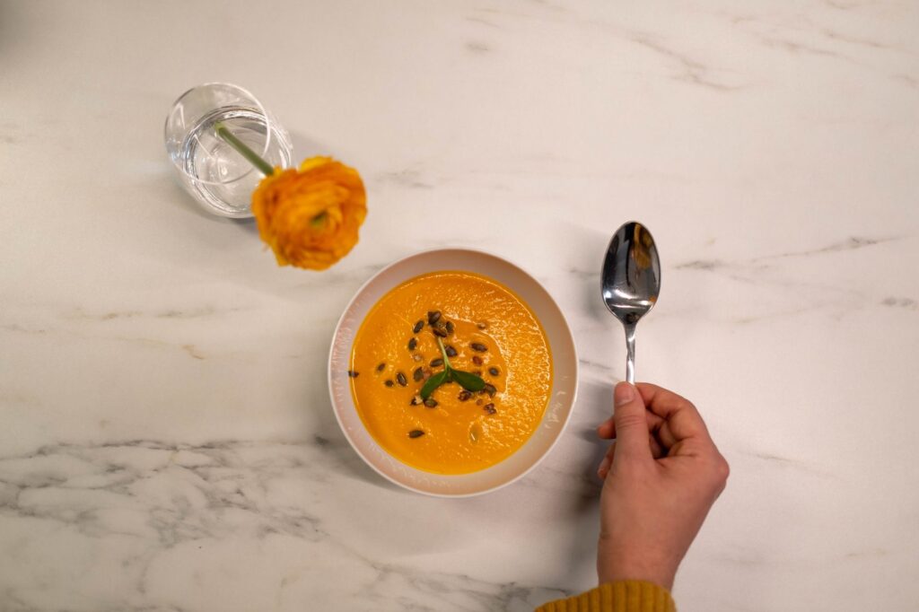Vue de dessus d'une soupe de potiron crémeuse garnie de graines et d'herbes, accompagnée d'une fleur jaune vif dans un vase en verre.