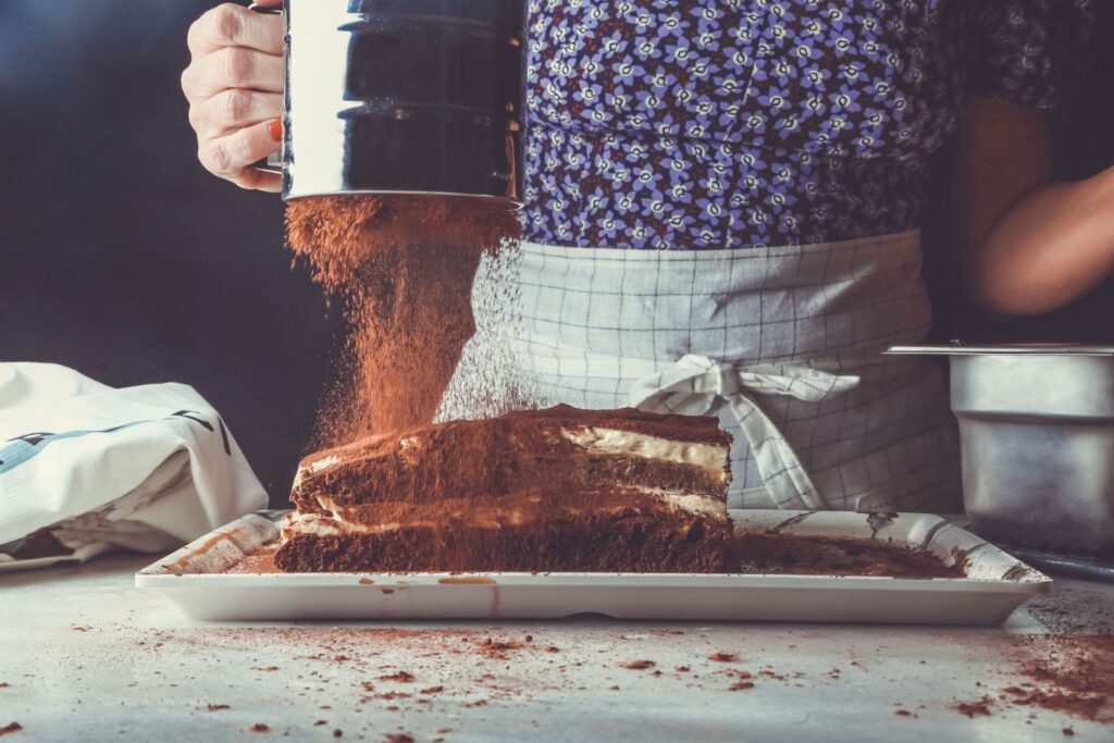 Une personne saupoudre du cacao sur un tiramisu crémeux dans une cuisine, mettant en avant la préparation de desserts faits maison.