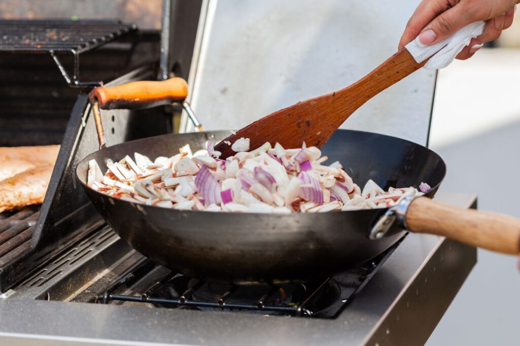 Gros plan sur une main cuisinant des champignons et des oignons dans une poêle en extérieur avec une spatule en bois.
