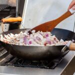 Gros plan sur une main cuisinant des champignons et des oignons dans une poêle en extérieur avec une spatule en bois.