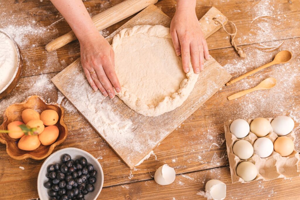 Mains d'une femme façonnant de la pâte sur une planche en bois entourée d'œufs et de myrtilles.
