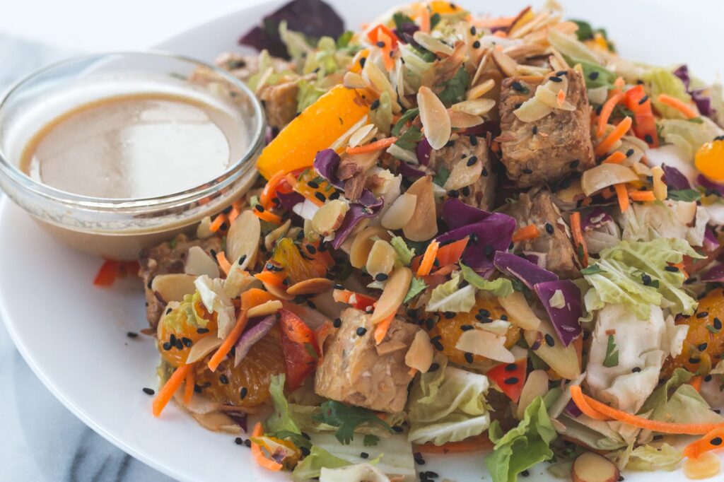 Une salade végétalienne vibrante avec du tempeh, des tranches d'orange et des amandes, servie avec une sauce.