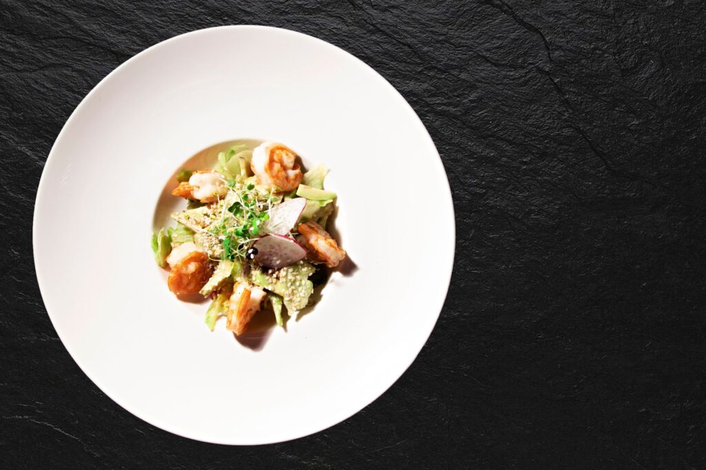 Vue aérienne d'une salade de crevettes et avocat fraîchement présentée sur une assiette blanche contre un fond texturé noir.