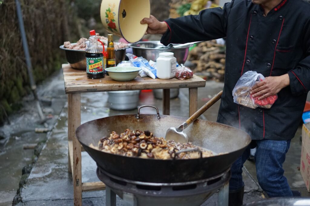 Chef remuant des ingrédients dans un wok en extérieur, mettant en valeur la cuisine asiatique. Comprend des bols d'ingrédients et des vêtements traditionnels.