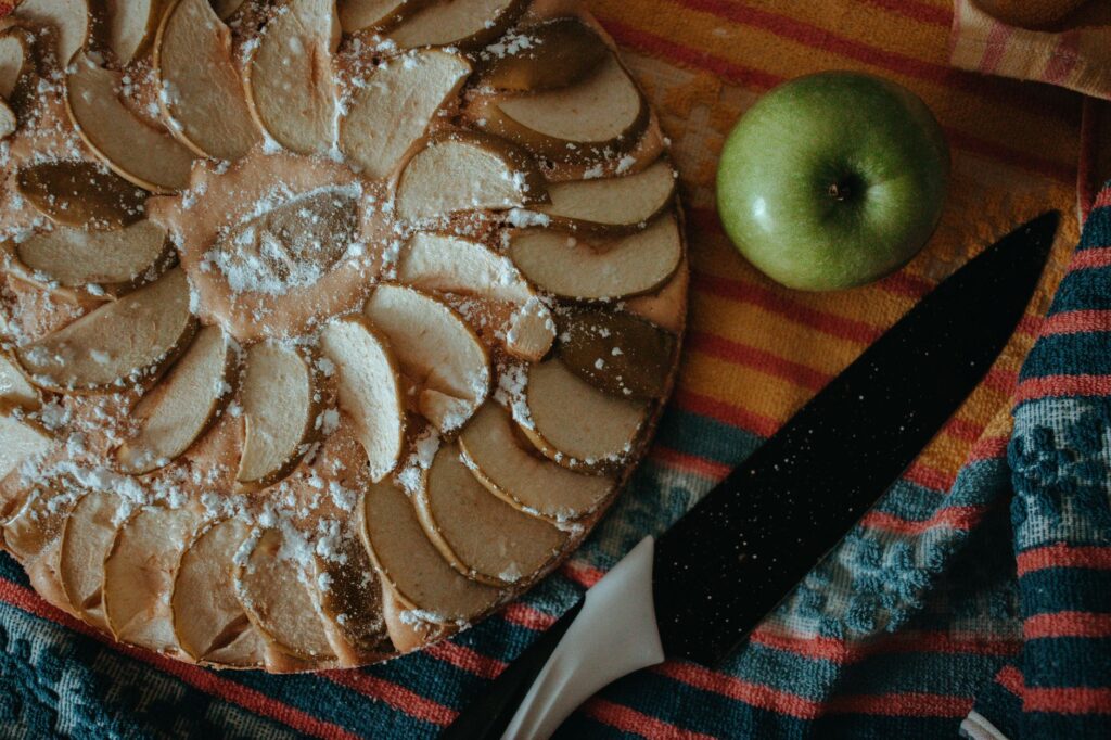 Tarte aux pommes savoureuse avec une pomme fraîche et un couteau sur un tissu coloré, vue de dessus.
