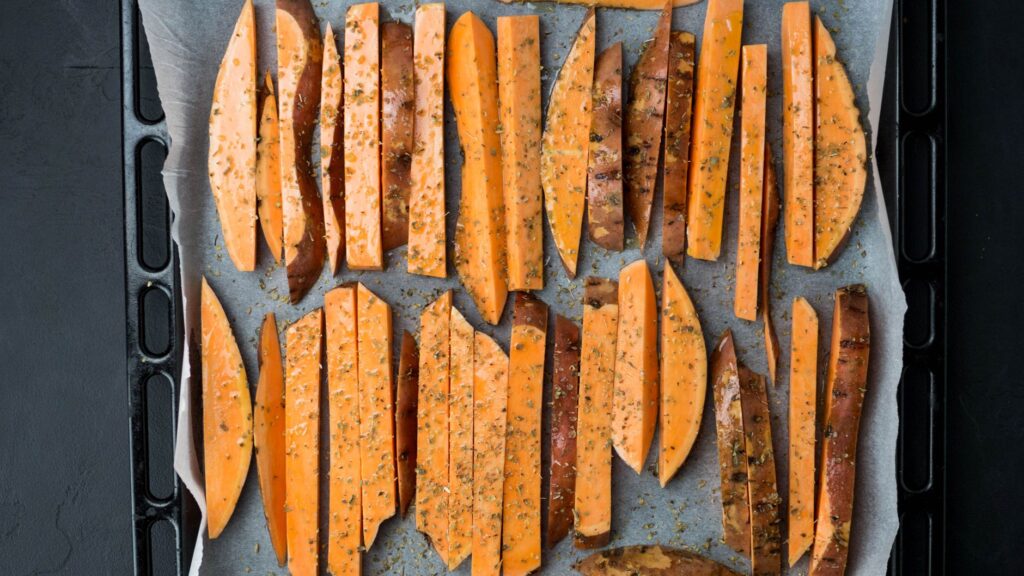 Gros plan de patates douces tranchées assaisonnées et disposées sur une plaque de cuisson avec du papier sulfurisé.